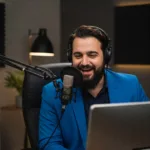 A comedy professional sits in front of his laptop speaking into a microphone, recording the latest instalment of his comedy podcast. Things to boost his comedian career!
